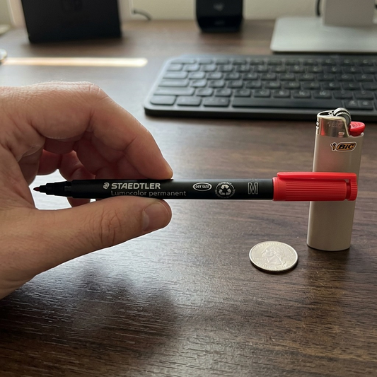 A photo-realistic scale comparison of a red STAEDTLER permanent marker with a person's hand.