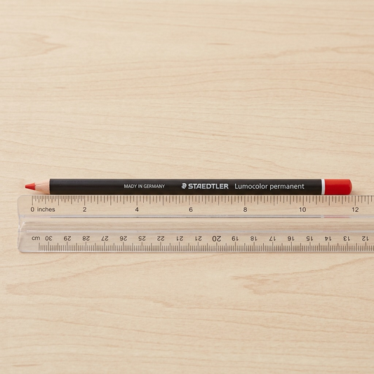 A close-up image showing a STAEDTLER Lumocolor permanent glasochrom dry marker placed alongside a transparent plastic 12-inch ruler on a light wood desk, offering a clear scale reference.