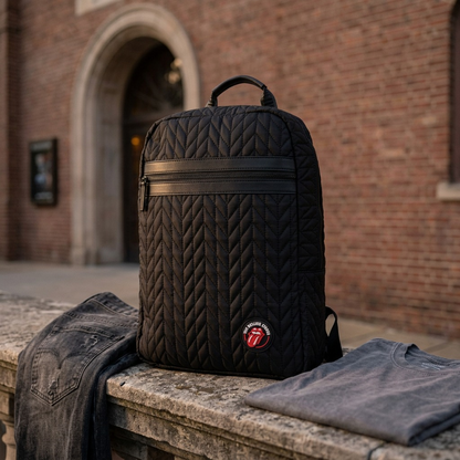 A black quilted Rolling Stones backpack with a leather trim rests on a weathered stone ledge outside a brick concert hall at dusk.