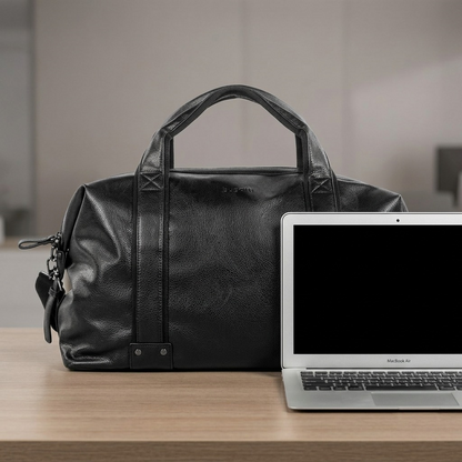 Bugatti Valentino black leather weekender bag next to a 13-inch laptop for size reference on a wooden desk.