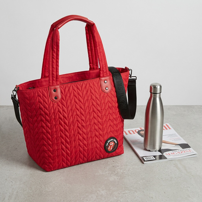 Product image of the red quilted The Rolling Stones tote bag shown sitting next to a reusable water bottle and a standard magazine, providing a clear visual representation of its physical dimensions.