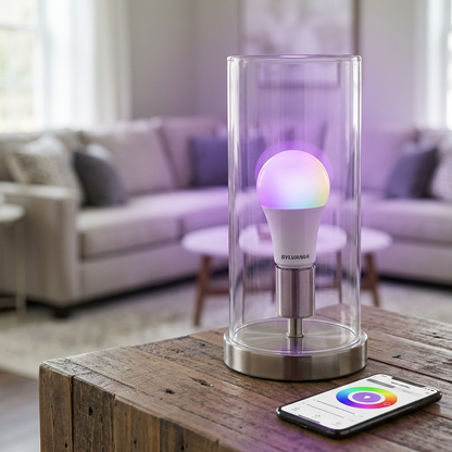 A SYLVANIA SMART+ WiFi A21 light bulb, accurately matching image_0.png, glows with a purple hue from its full color spectrum in an open table lamp socket. No packaging is present. Correct natural lighting and scale are used.