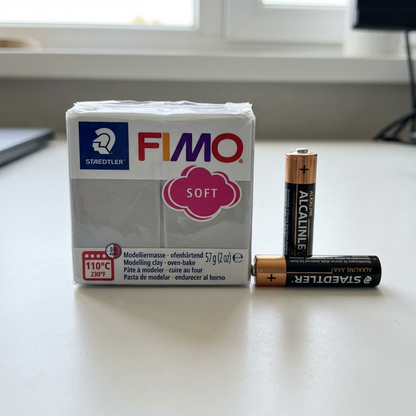 A full-frame photograph of a STAEDTLER Fimo Soft Dolphin Grey modeling clay package placed beside a standard AAA battery, showing its exact scale.