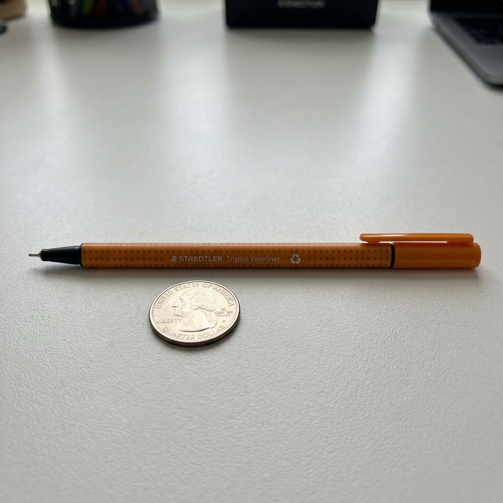 A close-up shot of the Staedtler Triplus fineliner beside an American quarter on a textured surface, showing relative size.