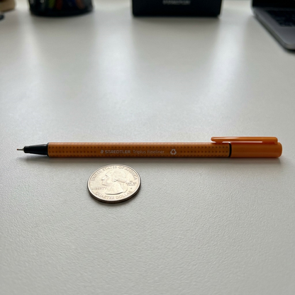 A close-up shot of the Staedtler Triplus fineliner beside an American quarter on a textured surface, showing relative size.