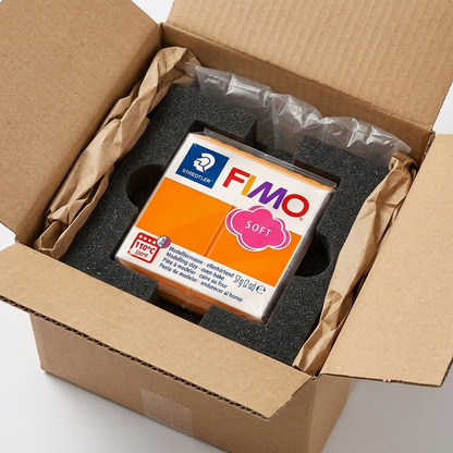 A photo of Fimo Soft tangerine modelling clay, in clear original packaging, set into custom-molded black foam within a partially open corrugated shipping box. The STAEDTLER Fimo labeling is clearly visible. The product is securely surrounded by protective padding, conveying safe delivery.