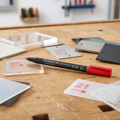 A Staedtler Lumocolor permanent red marker lies on a wooden desk next to sample material tags.
