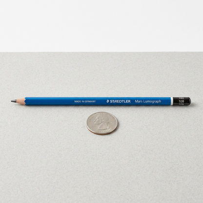 A photo of the Mars Lumograph 10B pencil horizontally on a desk beside a US Quarter for scale reference.