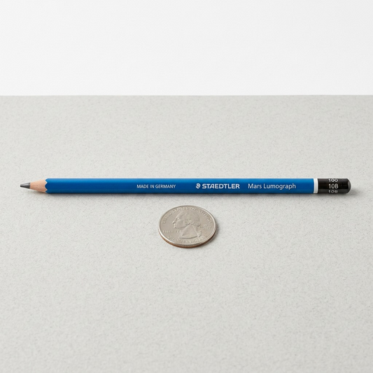 A photo of the Mars Lumograph 10B pencil horizontally on a desk beside a US Quarter for scale reference.