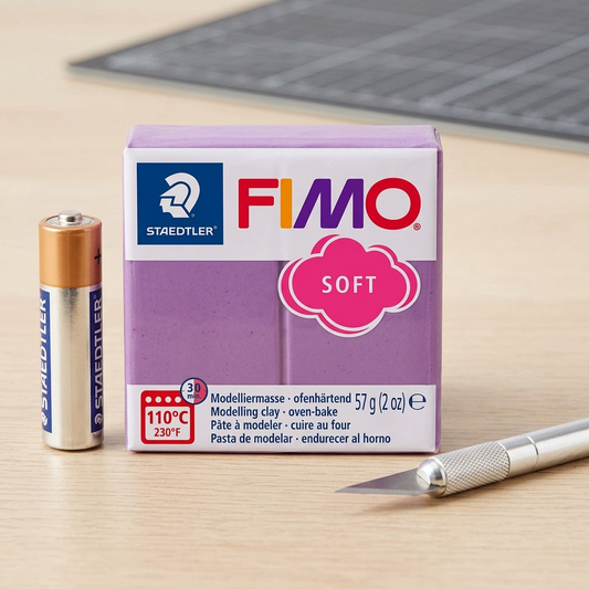 A lavender Fimo modeling clay block beside a AA battery and craft knife on a wooden desk.