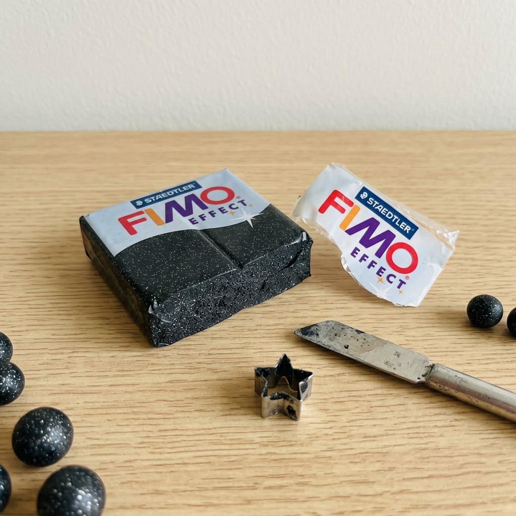 Lifestyle photo of STAEDTLER FIMO Effect Stardust glitter clay block unpeeled near a metallic jewelry-making hobby tool and a cut-out star shape on a white desktop.