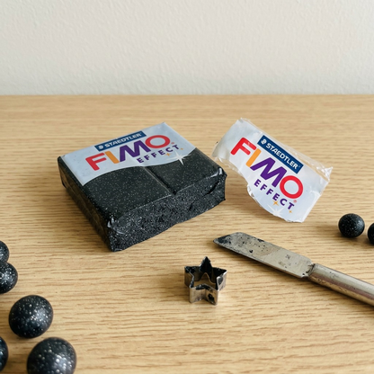 Lifestyle photo of STAEDTLER FIMO Effect Stardust glitter clay block unpeeled near a metallic jewelry-making hobby tool and a cut-out star shape on a white desktop.