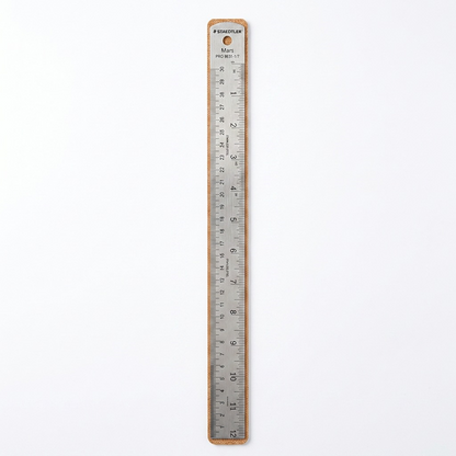 A premium, close-up photograph of the Staedtler Mars limited edition stainless steel ruler on a pure white background. The full length of the twelve-inch ruler is visible, showing the detailed imperial and metric measurements, the cork back, and the Staedtler branding at the top. The brushed metal texture is clear, and a subtle drop shadow grounds the object.