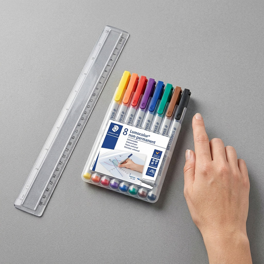 A STAEDTLER Lumocolor 8-pack is placed beside a 30 cm ruler on a minimalist grey desk surface, illustrating its size.