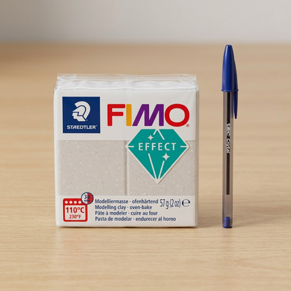 Scale reference photo of the 8010-002 Fimo Galaxy White clay block, and its clear plastic packaging, shown next to a standard plastic ballpoint pen, resting on a clean wooden desk surface. Studio lighting.