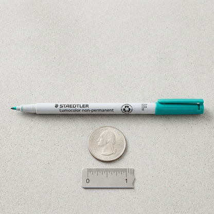 A STAEDTLER fine green pen next to a U.S. quarter and a small metal ruler on a grey surface.