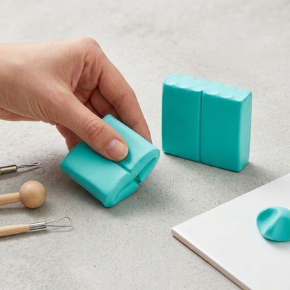 A flatlay photograph on a neutral grey concrete surface shows a hand holding the Fimo Soft Peppermint clay, actively kneading and softening it, surrounded by simple sculpting tools, illustrating its use.