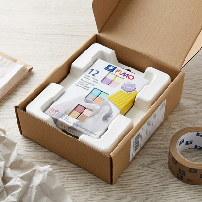 A high-angle photo shows a box of Fimo Soft Pastel 12 Color Pack being carefully unboxed from a well-padded shipping carton on a wood surface. The retail packaging is intact and clear within a form-fitting paper pulp insert. Padded mailer and crumpled paper packing material are next to the brown shipping box with an generic label. Natural daylight. Accurate product details.