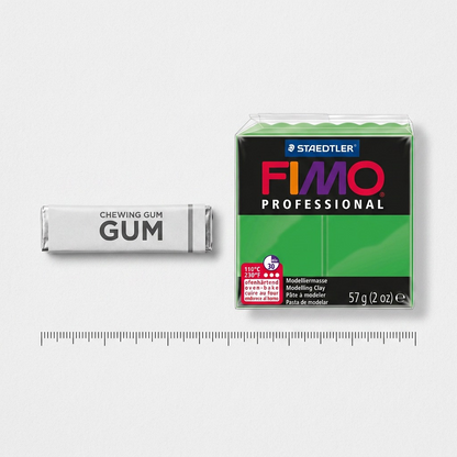 A photograph showing a standard stick-of-gum package placed immediately to the left of the STAEDTLER Fimo Professional True Green 57g (2 oz) clay package, directly comparing their sizes on a minimalist surface to provide a clear scale reference, set against a clean, neutral studio background with soft, natural lighting.