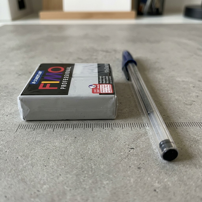 FIMO Professional Dolphin Gray clay brick shown to scale next to a blue Bic ballpoint pen on a concrete surface with measurement markings.