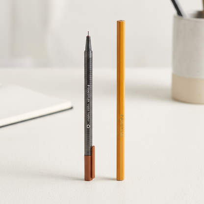A close-up photograph of the STAEDTLER triplus fineliner next to a standard wooden pencil on a desk.