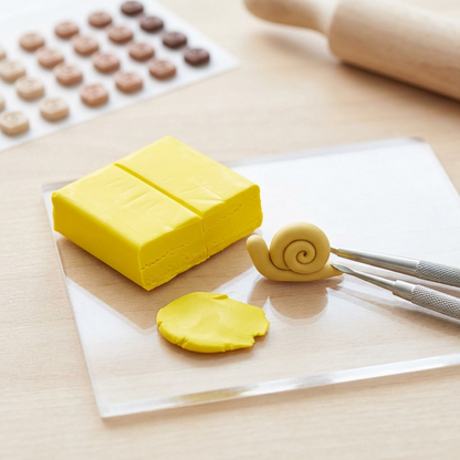 A natural daylight photo of one opened STAEDTLER FIMO Professional True Yellow 57g block and one raw, unbaked miniature snail figure made from it, resting on a clean, light wooden workshop desk.