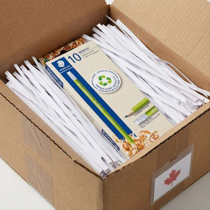 A close-up photograph of a STAEDTLER Wopex pencil pack, identical to the reference, nestled safely within a brown cardboard shipping box overflowing with clean white protective paper shredding, showing the box corners reinforced with brown paper tape.