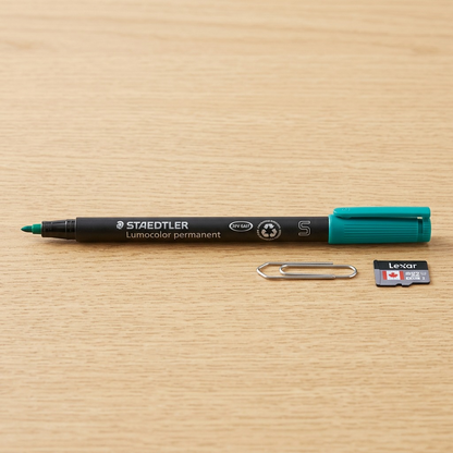 A detailed photo of the STAEDTLER permanent marker with its distinct teal cap, placed horizontally alongside a standard metal paperclip on a desk surface, showcasing the pen's compact fine point and true size. The smooth matte black body and all branding details from the reference are visible. The natural wood grain background provides a clear scale comparison.