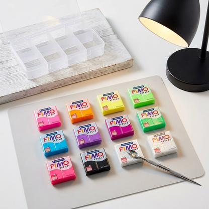An overhead photograph displays twelve rectangular blocks of vibrant neon FIMO Effect polymer clay resting neatly in rows on a light gray craft mat. A steel-blade sculpting tool is positioned near the corner of one white block. To the upper left, an open, empty clear plastic organizer sits on a whitewashed wooden table, next to a black desk lamp. The scene is illuminated by soft, natural daylight.