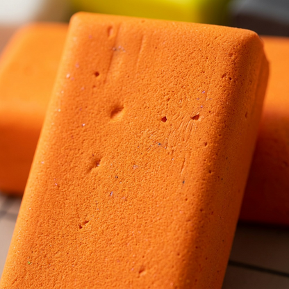 An extreme macro photograph of the vibrant, granular texture and soft, malleable surface of a STAEDTLER Fimo Effect Neon clay block, showing the intense neon orange pigment under precise product-led lighting.