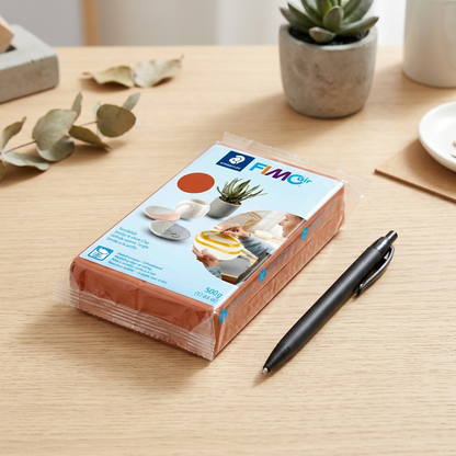 Fimoair 500g clay block sits on a wooden desk next to a pen for a clear size comparison.