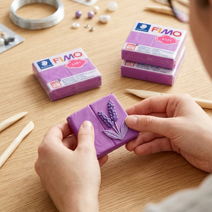 A person molds a piece of purple FIMO Soft modelling clay on a wooden table.