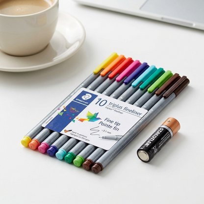 A close-up image showing a STAEDTLER triplus fins pen with cap on, as if taken from the canonical image_0.png set, sitting perfectly parallel to and touching the top-long-edge of a standard AA battery on a light grey wooden desk surface. Realistic soft studio lighting highlights both items.