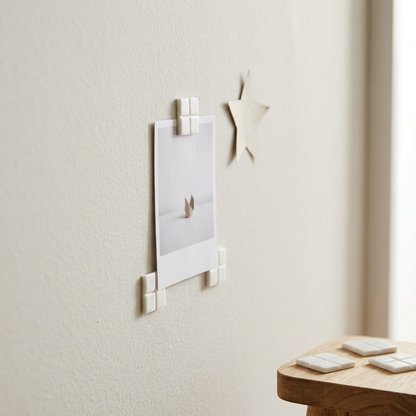 A close-up photograph of discrete, small white square adhesive pads from the UHU tac deco pack holding a printed photo to a minimalist plaster wall. The lighting is soft and natural.