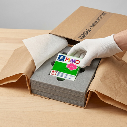 A premium, close-up photo of the STAEDTLER Fimo Soft Tropical Green clay block, securely nestled within a custom-fit, protective foam insert inside a sturdy, realistic cardboard shipping box, showcasing reliable, damage-free delivery.