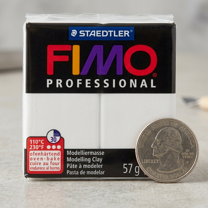 An eye-level product shot of the STAEDTLER FIMO Professional White 57g block, as referenced with all its specific multi-colored logos and details, sitting next to a standard American Quarter coin on a light wood desk.