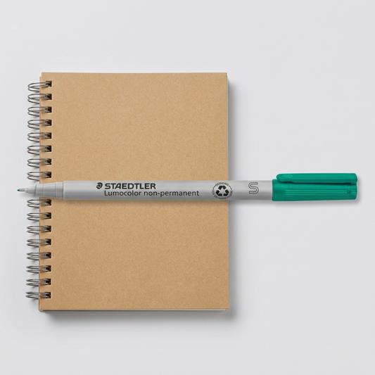 The STAEDTLER Lumocolor non-permanent marker from the reference image, placed beside a brown coil-bound A5 notebook for scale perspective, on a clean studio desk surface. Both objects are rendered in true proportion.