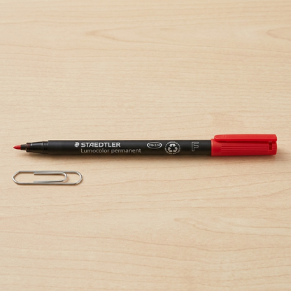 A photo of the red STAEDTLER permanent marker next to a standard metal paperclip on a desk surface.