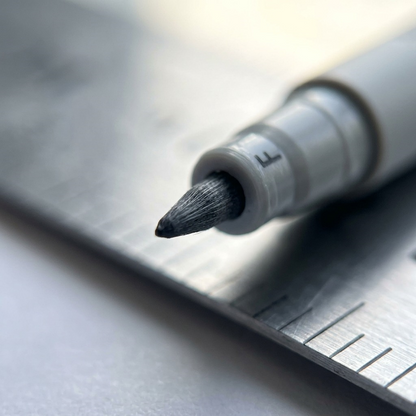 An extreme close-up photograph of a STAEDTLER Lumocolor non-permanent pen fine (F) tip with an 0.6mm fiber, set against a blurred gray background.