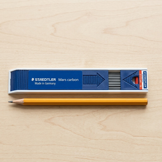 A realistic visual comparison showing a single case of STAEDTLER Mars Carbon 2mm Carbon Leads (200-2H-03) resting beside a standard wooden hexagonal pencil for an accurate scale reference.