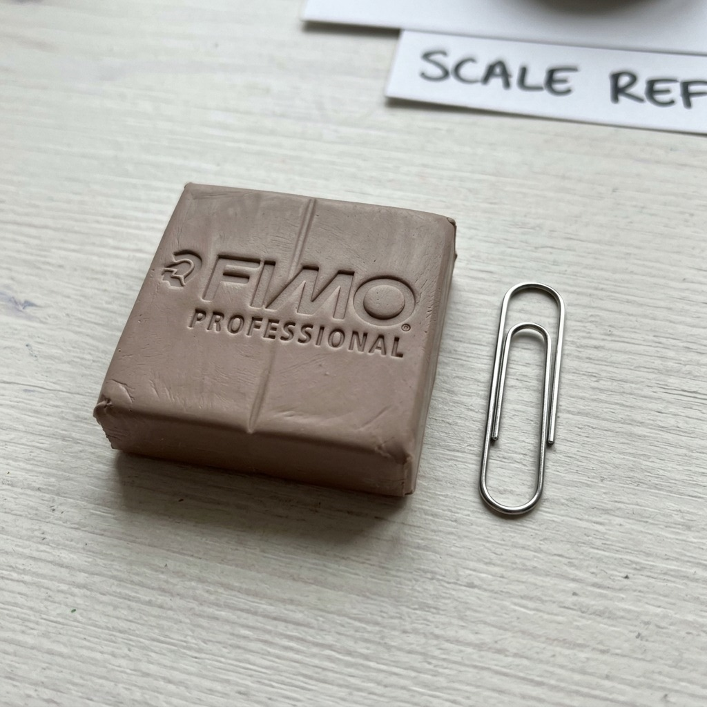 A photograph showing the STAEDTLER Fimo Professional Taupe 57g clay block sitting next to a common plastic paperclip on a desk for size comparison.