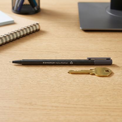 A STAEDTLER triplus Fineliner 334-8-A7, dark grey, resting precisely beside a standard house key on a clean desk.