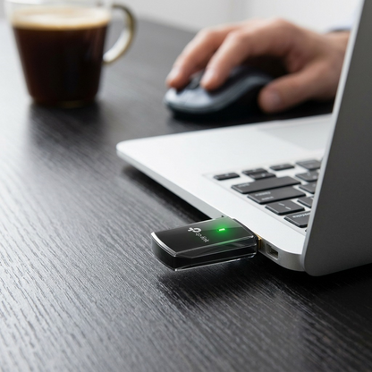 A close-up photograph of a laptop workspace featuring the TP-Link AC600 Wireless USB Adapter plugged into a port on the laptop's side, with the green indicator light visible.