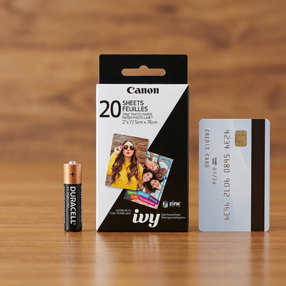 A ZINK paper pack sits beside an standard AA battery and a credit card on a wooden surface for size.