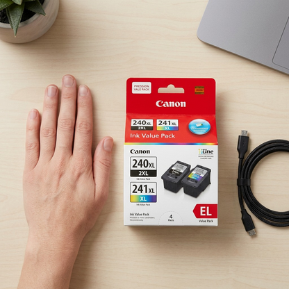 A close-up photograph of the Canon 5206B020 ink value pack box placed on a wooden desk next to a standard human hand, illustrating its physical scale. The shot includes a coiled micro-USB cable as an additional size reference.