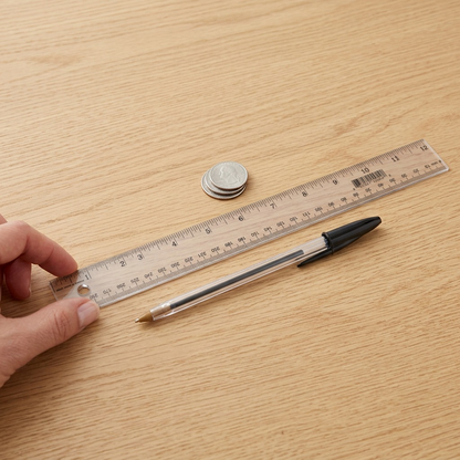 A clear photo of the 12-inch Westcott transparent ruler from image_0.png, resting on a wooden desk next to a human hand holding a standard pen, showing its length clearly for scale.